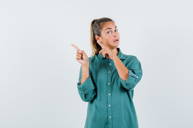 Mujer joven apuntando hacia la izquierda con los dedos índices en blusa verde y mirando serio