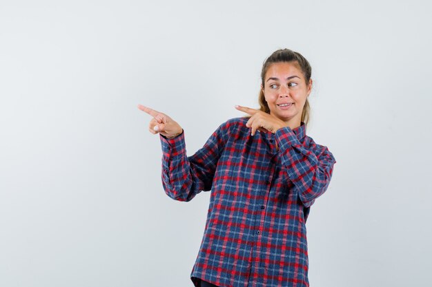 Mujer joven apuntando hacia la izquierda con los dedos índices en camisa a cuadros y luciendo bastante