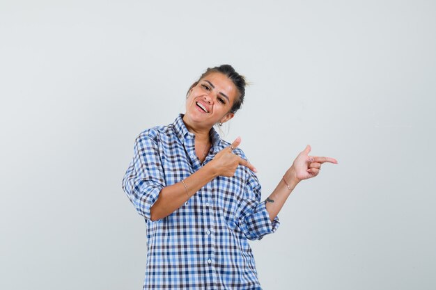 Mujer joven apuntando a un lado en camisa a cuadros y mirando alegre.