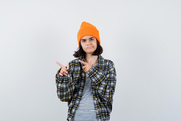 Mujer joven apuntando hacia el lado con camisa a cuadros sombrero naranja mirando infeliz