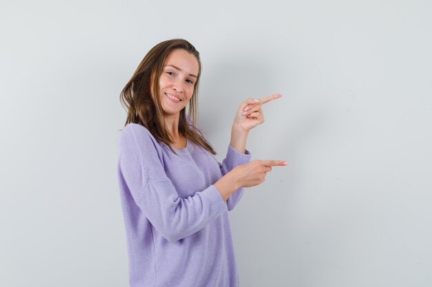 Mujer joven apuntando hacia el lado izquierdo en blusa lila y mirando satisfecho. vista frontal.