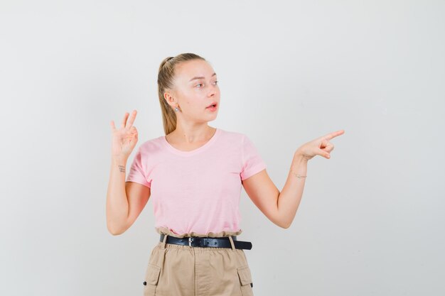 Mujer joven apuntando a un lado, mostrando el signo de ok en camiseta, pantalones y mirando enfocado. vista frontal.