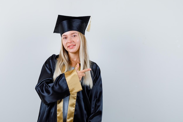 Mujer joven apuntando a un lado en uniforme de posgrado y mirando alegre.