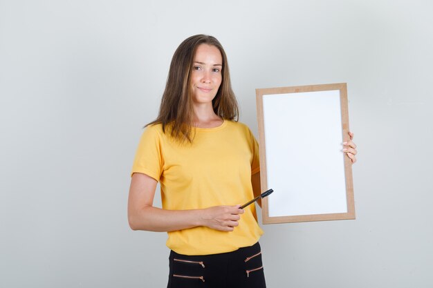 Mujer joven apuntando con lápiz en la pizarra blanca en camiseta, pantalón y mirando contento