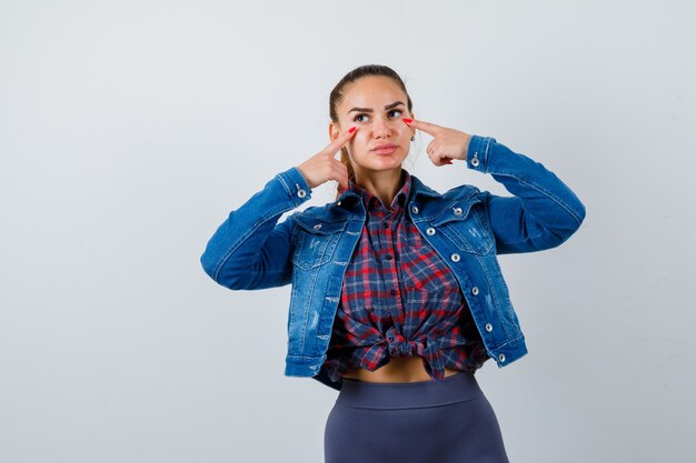 Mujer joven apuntando a los ojos con los dedos en camisa a cuadros, chaqueta de jean y mirando nostálgico, vista frontal.