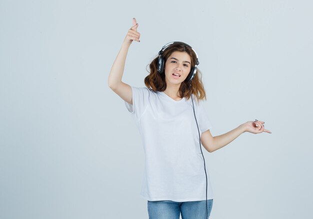 Mujer joven apuntando hacia otro lado mientras disfruta de la música con auriculares en camiseta blanca, jeans y mirando feliz, vista frontal.