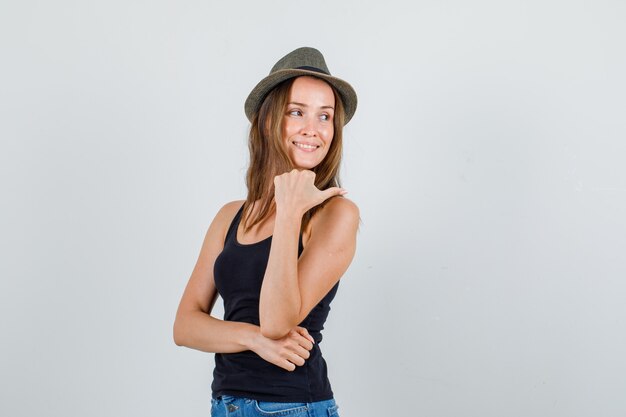 Mujer joven apuntando con el pulgar mientras mira hacia atrás en camiseta, pantalones cortos, sombrero y mirando contento