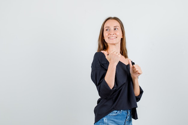 Foto gratuita mujer joven apuntando los pulgares hacia atrás en camisa, pantalones cortos y mirando alegre.