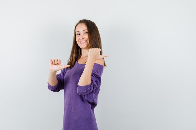 Mujer joven apuntando los pulgares hacia atrás en camisa violeta y mirando feliz. vista frontal.