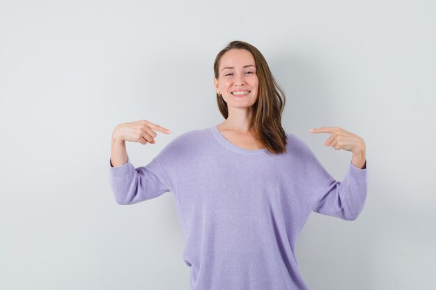 Mujer joven apuntando a sí misma en blusa lila y luciendo optimista. vista frontal.