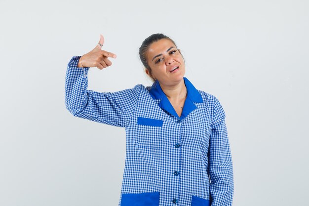 Mujer joven apuntando a sí misma con el dedo índice en camisa de pijama de cuadros azules y mirando bonita, vista frontal.