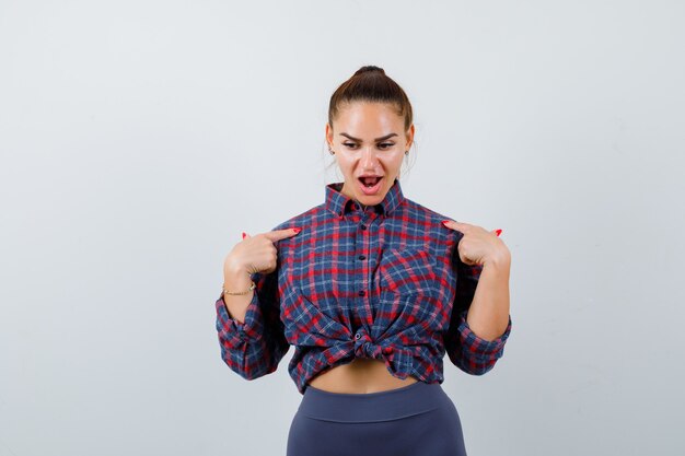 Mujer joven apuntando a sí misma mientras mira hacia abajo en camisa a cuadros, pantalones y mirando perplejo. vista frontal.