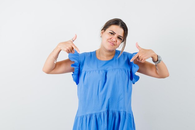 Mujer joven apuntando a sí misma en vestido azul y luciendo orgullosa