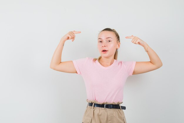 Mujer joven apuntando a su cabeza en camiseta, pantalón y mirando sensible, vista frontal.