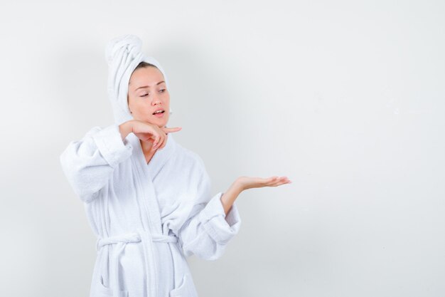 Mujer joven apuntando a su palma extendida a un lado en bata de baño blanca, toalla y mirando confiada, vista frontal.