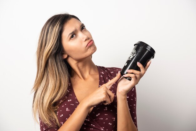 Mujer joven apuntando a una taza de bebida en la pared blanca.