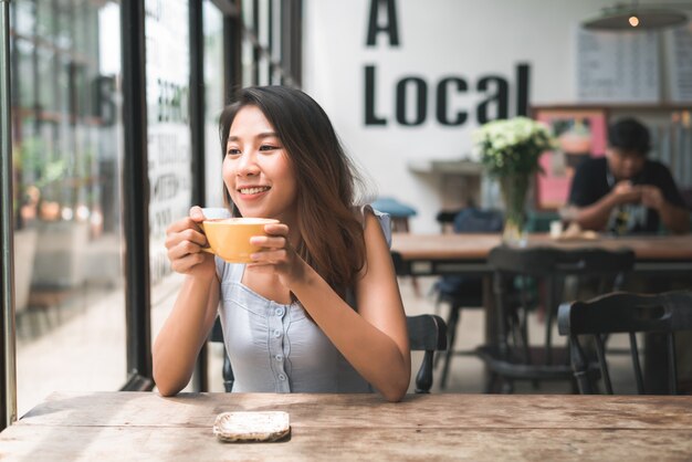 Mujer joven asiática alegre que bebe el café o el té caliente que lo goza mientras que se sienta en café