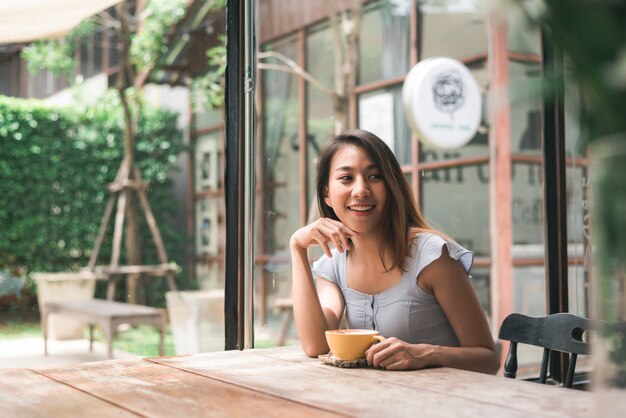 Mujer joven asiática alegre que bebe el café o el té caliente que lo goza mientras que se sienta en café