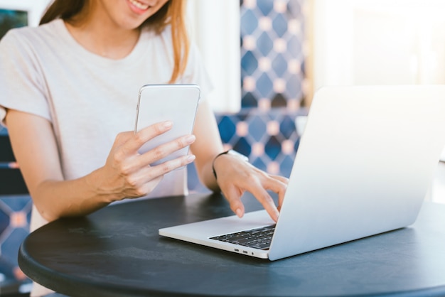 Mujer joven asiática alegre que se sienta en café de consumición del café y que usa el smartphone para hablar