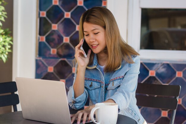 Mujer joven asiática alegre que se sienta en café de consumición del café y que usa el smartphone para hablar