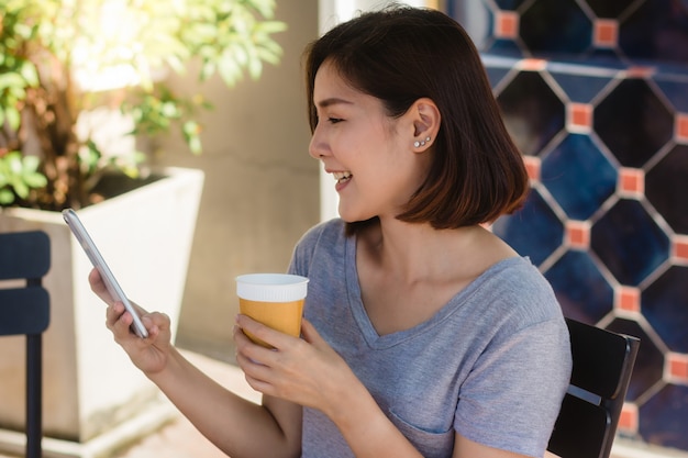 Mujer joven asiática alegre que se sienta en café de consumición del café y que usa el smartphone para hablar