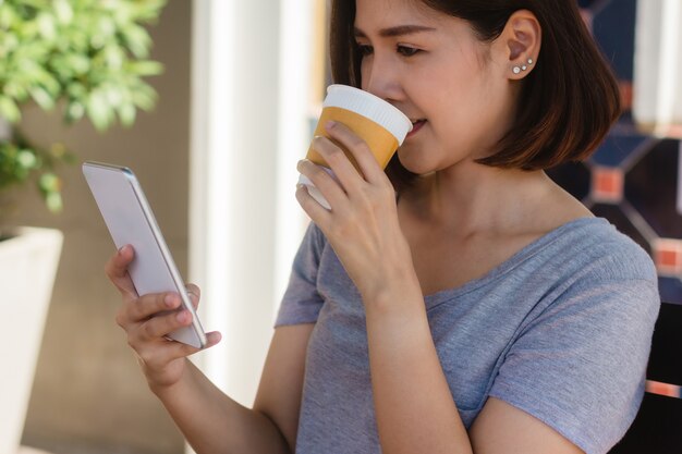 Mujer joven asiática alegre que se sienta en café de consumición del café y que usa el smartphone para hablar