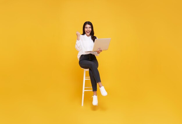 Mujer joven asiática feliz sonriendo mientras ella usa una computadora portátil sentada en una silla blanca