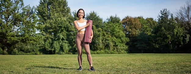 Mujer joven asiática segura de sí misma y deportiva de pie en el parque con estera de goma vestida de deportista sonriendo