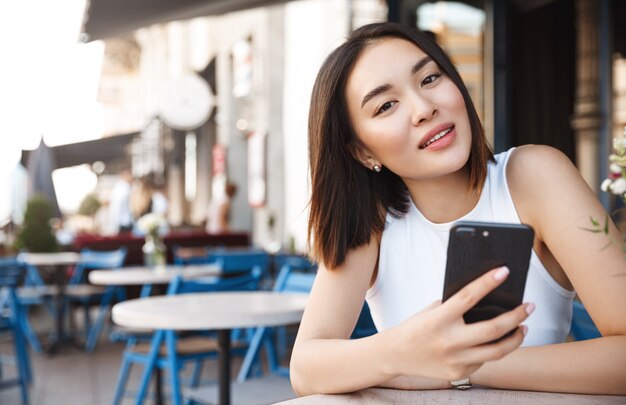Mujer joven asiática sentada en la cafetería con teléfono móvil, mirando a la cámara soñadora