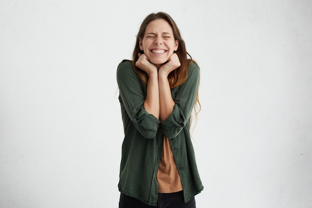 Foto gratuita mujer joven asombrada con cabello lacio oscuro cerrando los ojos y apretando los puños con placer teniendo una amplia sonrisa siendo feliz