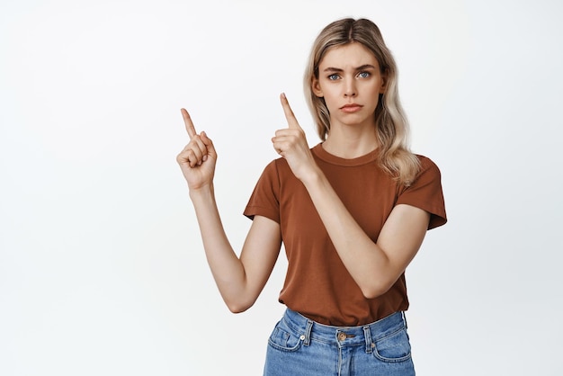 Mujer joven de aspecto serio que señala con el dedo en la esquina superior izquierda mostrando algo importante haciendo un anuncio de pie contra el fondo blanco