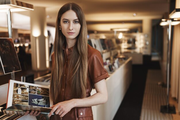 Mujer joven de aspecto serio recogiendo revistas de diseño en la tienda vintage, de pie con un elegante vestido marrón.