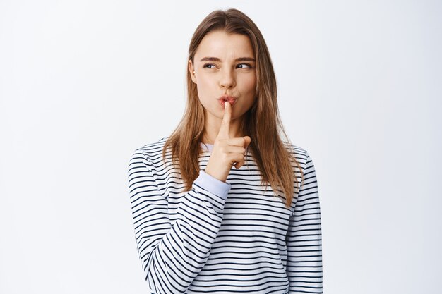 Mujer joven astuta que hace silencio, hace un letrero de silencio en los labios y mira a la izquierda pensativa, tiene un secreto, pide estar en silencio, hace un letrero tabú sobre una pared blanca