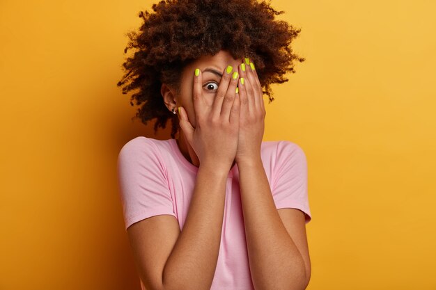 Una mujer joven asustada y asustada se asoma a través de los dedos, esconde la cara, mira con los ojos muy abiertos algo espeluznante, usa una camiseta informal, se encuentra alarmada contra la pared amarilla, está preocupada