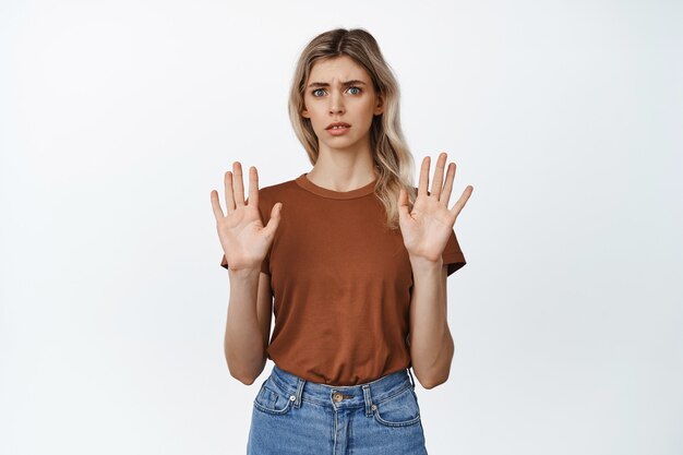 Mujer joven asustada levantando las manos vacías, rechazando algo, rechazando la oferta y diciendo que no, de pie en camiseta y jeans en blanco.