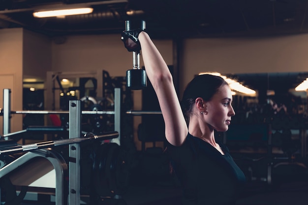 Mujer joven atlética que hace ejercicio del tríceps con pesa de gimnasia en centro de aptitud