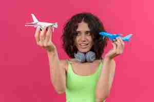 Foto gratuita una mujer joven y atractiva alegre con el pelo corto en la parte superior de la cosecha verde en auriculares sosteniendo aviones de juguete blanco y azul