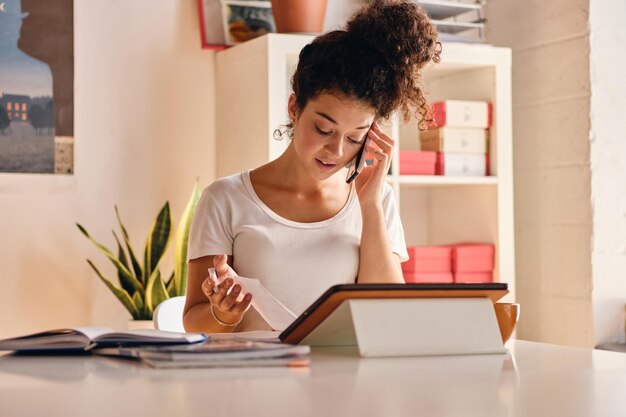 Mujer joven y atractiva con cabello rizado oscuro sentada en la mesa con un cuaderno y una tableta hablando cuidadosamente por teléfono celular y estudiando en un hogar moderno y acogedor