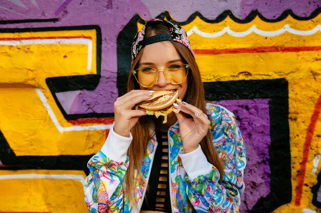 Mujer joven y atractiva, comiendo una sabrosa hamburguesa. Vestido con chaqueta y gorro coloridos