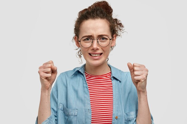 La mujer joven y atractiva cree firmemente en el éxito, mantiene las manos con los puños cerrados, tiene una expresión facial deseable, vestida con ropa a rayas, aislada en la pared blanca. Concepto de lenguaje corporal