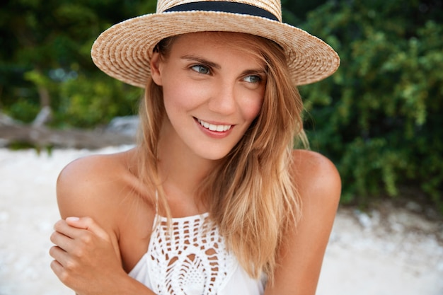 Mujer joven atractiva con expresión positiva de ensueño, vestida con sombrero de moda y vestido blanco de verano, mira pensativamente a la distancia, planea algo para el día siguiente, tiene un buen descanso en el extranjero