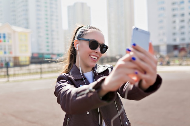 Mujer joven atractiva en gafas de sol negras y chaqueta de cuero haciendo selfie