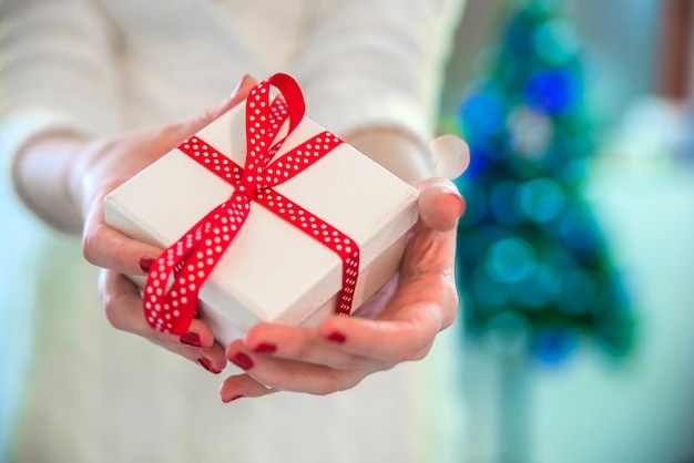 Mujer joven atractiva hermosa en el suéter blanco que se sienta al lado del árbol de navidad blanco, sosteniendo el presente. Foto de navidad