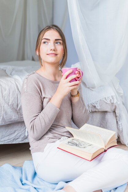 Mujer joven atractiva con libro sobre vuelta sosteniendo la taza de café mirando a otro lado