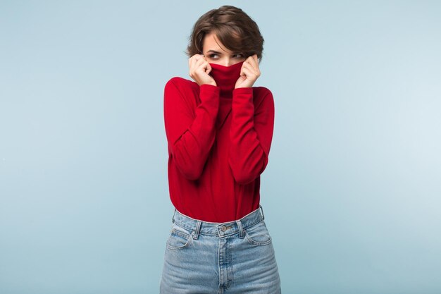 Una mujer joven y atractiva con el pelo corto y oscuro escondiéndose en un suéter rojo mientras mira cuidadosamente a un lado sobre el fondo azul