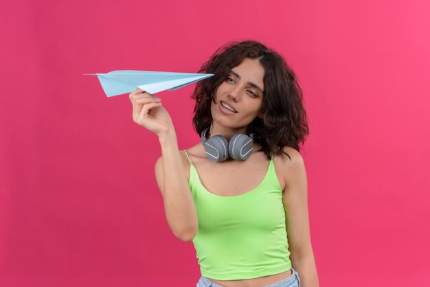 Una mujer joven y atractiva con el pelo corto en verde crop top en auriculares mirando un avión de papel azul