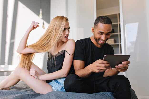 Mujer joven atractiva con el pelo largo y rubio en pantalones cortos está enojada con el chico guapo alegre jugando en la tableta cerca de la cama en su casa en un apartamento moderno. Divirtiéndose, relación, amor, pareja divertida