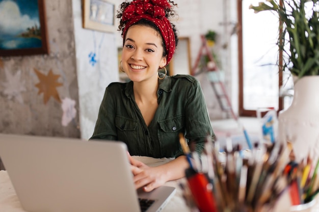 Foto gratuita mujer joven y atractiva con el pelo rizado oscuro sentada en la mesa con una laptop mirando soñadoramente a la cámara mientras pasa tiempo en un taller moderno y acogedor con grandes ventanas