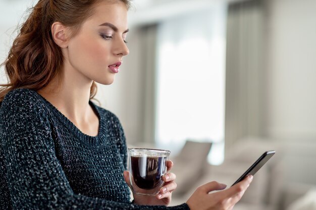 Mujer joven atractiva que mira su teléfono inteligente en casa. Mensaje de tipos de mujer en su teléfono inteligente.