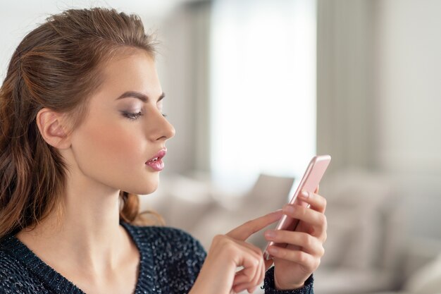 Mujer joven atractiva que mira su teléfono inteligente en casa. Mensaje de tipos de mujer en su teléfono inteligente.